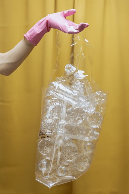 A person wearing a pink rubber glove holds a clear plastic bag filled with various empty plastic bottles and containers, some with caps and labels still visible. The plastic items are of different shapes and sizes, and are loosely stacked inside the bag. The background features a solid yellow curtain, providing a neutral backdrop that emphasizes the cleanliness and transparency of the plastic waste. The scene suggests an activity related to recycling or collection within a waste management context, possibly part of a private waste collection service such as Rubbish Removal Waterloo, which specializes in alternative waste handling including rubbish clearance and environmentally conscious disposal. The image highlights the importance of proper sorting and disposal of plastic waste in a waste management process, supporting the broader theme of rubbish removal and recyclable waste separation for domestic or commercial clearance, particularly in urban environments.