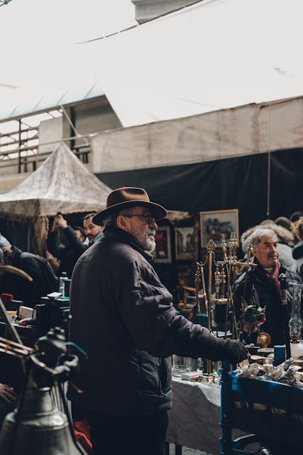 An older man with a grey beard, wearing a brown wide-brimmed hat and dark clothing, stands at an outdoor market stall in a busy marketplace setting. The stall displays various vintage items and collectibles, with a focus on metal and glass objects, arranged on a table covered with a white cloth. Behind him, several other traders and shoppers are visible, along with framed artworks hanging on a wall and a tent overhead providing shelter from the weather. The environment appears to be bustling, with multiple people engaged in conversation and browsing. The scene is illuminated by natural daylight, enhancing the details of the textured surfaces and diverse colors of the items for sale. This setting reflects a typical marketplace environment where independent traders, such as those managed by Rubbish Removal Waterloo, might operate, and subtly aligns with the theme of private disposal and on-site clearance of unwanted goods or waste material.