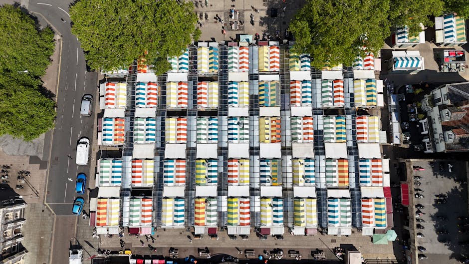 An aerial view of a market square filled with numerous vendor stalls arranged in a grid pattern, each covered by colorful striped canopy tents in red, yellow, blue, and green. The stalls are closely packed, with narrow pathways between them, and appear to be set up on a paved surface. In the surrounding area, trees with lush green foliage partially obscure the edges of the market, and nearby streets show parked cars and pedestrians. The scene suggests a bustling outdoor market environment, where independent vendors may handle on-site waste management, aligning with the context of rubbish removal services for markets and stalls. The image emphasizes the organized coverage of multiple stalls, highlighting the need for efficient rubbish collection and disposal in such busy public spaces, which companies like Rubbish Removal Waterloo may service through their waste removal expertise.