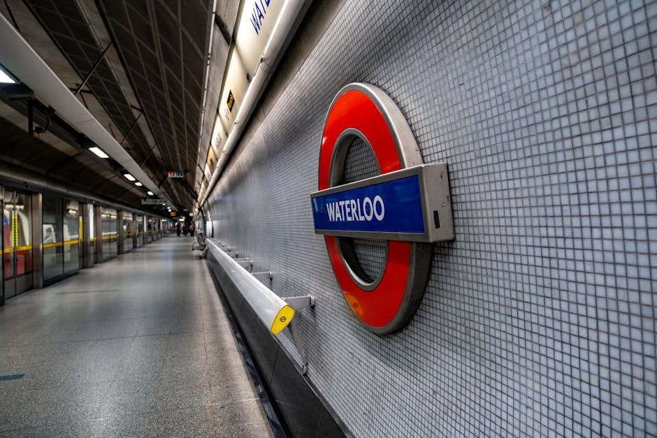 An underground station platform at Waterloo features a large, circular London Underground roundel sign mounted on a wall tiled with small, light grey square tiles. The sign displays the station name 'Waterloo' on a blue rectangular background with white text, partially extending from the roundel's red ring. The platform is empty with a smooth, polished grey floor, and a yellow safety line runs along the edge next to a white safety barrier. Overhead lighting illuminates the scene, reflecting off the tiled wall and the clean surface of the platform. On the left side, glass partition walls separate the platform from the tracks, with metal framing and reflections visible on the glass. The overall environment appears modern and well-maintained, typical of urban railway stations, supporting the context of efficient public transportation hubs that complement private waste management and rubbish removal services in the area.
